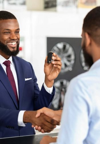 Black Salesman And Male Customer Shaking Hands After Successful Deal In Dealership Center, Friendly Salesperson Giving Car Keys To Client, Greeting With Purchase In Auto Showroom, Selective Focus